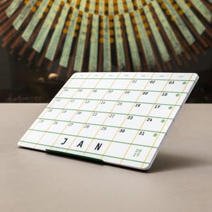Puede incluir: Un calendario de escritorio blanco para enero de 2027 con una cuadrícula de líneas verdes y amarillas. El calendario muestra los días de la semana en la parte superior, las fechas del 01 al 31 y el texto JAN 2027. Se apoya en un soporte de metal verde oscuro.