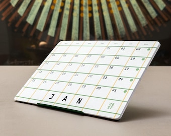 May include: A white desk calendar for January 2027 featuring a grid with green and yellow lines. The calendar displays the days of the week at the top, dates from 01 to 31, and the text JAN 2027. It is supported by a dark green metal stand.