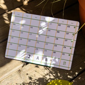 May include: A rectangular desk calendar for January 2027 featuring a white background with a grid of thin green and yellow lines. The calendar displays the days of the week, dates from 01 to 31, and the text JAN 2027 in bold black lettering.