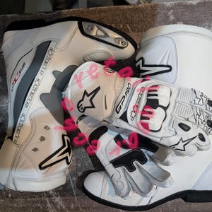 Könnte beinhalten: Weiße Motorradstiefel und -handschuhe. Die Stiefel haben schwarze Akzente und graue Streifen, wobei der Markenname sichtbar ist. Die Handschuhe sind weiß mit schwarzen und rosa Details, und der Text "GP Pro" ist sichtbar.