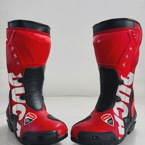 Puede incluir: Par de botas de moto rojas y negras con el logo "Ducati" en blanco. Las botas tienen una parte superior de cuero rojo perforado, detalles en negro y una suela negra. Diseñadas para motociclismo.