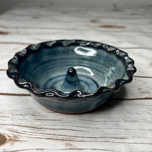 May include: A round ceramic ring dish with a blue and grey glazed finish. The dish features a ruffled, scalloped edge and a small, pointed cone in the center for holding rings. The interior surface shows subtle circular ridges from the pottery process.