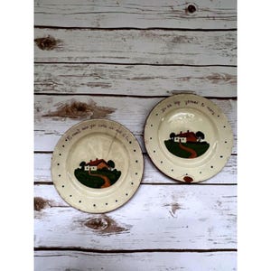 May include: Two off-white ceramic plates with a hand-painted house scene. Each plate features a red-roofed house, a green hill, and a brown road. The plates have black dot borders and text in a script font.