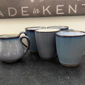 May include: A collection of ceramic mugs and a creamer with a blue glaze and brown trim. The mugs have handles and a rounded shape. The creamer has a handle and a spout. The text "MADE in KENT" is visible in the background.