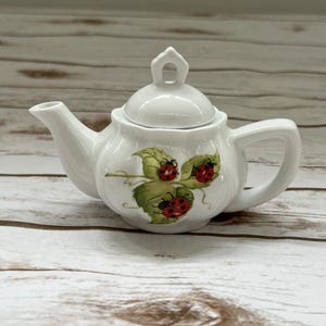 May include: A small, white ceramic teapot with a lid and handle. The teapot is decorated with a design of green leaves and red ladybugs. The spout and handle are curved, and the lid has a small loop for easy removal. The teapot is likely for decorative use.