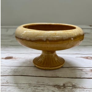 May include: A ceramic pedestal bowl featuring a glossy finish with a gradient of deep amber at the base transitioning to a lighter cream color along the wide, rounded rim. The bowl is supported by a short, fluted stem and a circular base.