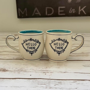 May include: Two cream-colored ceramic mugs with a blue interior and a decorative handle. Each mug features the text "HELLO THERE" in a black ornate frame. The mugs are set on a light-colored wooden surface.
