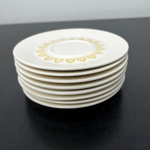May include: A stack of off-white saucers with a gold floral design around the center. The saucers have a ridged edge and are stacked on a dark surface. The saucers are likely for a tea or coffee set.