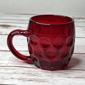 May include: A ruby red glass mug with a handle. The mug has a textured pattern of raised circles on the sides. The mug is sitting on a white wooden surface.