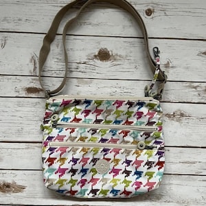 May include: A white crossbody bag with a colorful houndstooth pattern in shades of pink, green, blue, and yellow. The bag has three zippered compartments and a tan adjustable strap. A small, round logo is visible on the front.
