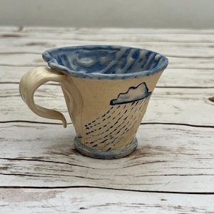 May include: A ceramic mug with a cream-colored exterior featuring a hand-painted blue cloud and rain pattern. The interior rim is glazed in blue with white drip accents. It has a curved, cream-colored handle and a flared, uneven top edge.