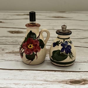 May include: Two ceramic bottles with floral designs. One bottle has a handle and a red flower design, the other has a blue flower design. Both bottles have decorative stoppers and are on a wooden surface.