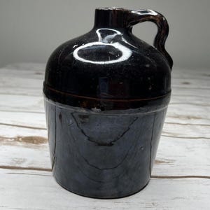 May include: A dark brown ceramic jug with a handle. The jug has a rounded top and a cylindrical base. The surface has a glossy finish. The jug is sitting on a light-colored wooden surface.