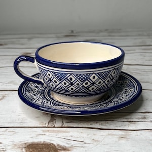May include: A ceramic teacup and matching saucer featuring a hand-painted dark blue geometric pattern on a cream-colored background. The design consists of repeating diamonds, cross-hatching, and small dots, with a solid blue rim and handle.