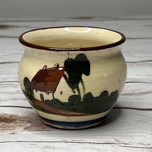 May include: A small, round ceramic bowl with a cream-colored exterior and a dark brown rim. The bowl features a hand-painted landscape scene with a house, trees, and a path in shades of brown, green, and blue. The bowl is approximately 3 inches in diameter.