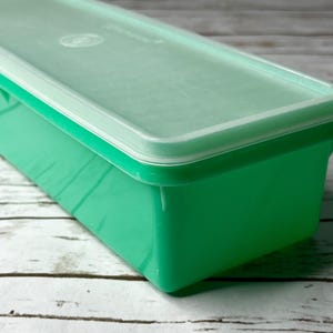 May include: A rectangular storage container with a vibrant green base and a translucent, semi-opaque lid. The container features a smooth, solid-colored plastic construction with a secure-fitting top, designed for organizing or storing various items.