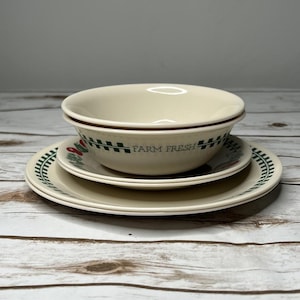 May include: A cream-colored ceramic bowl and two plates are stacked. The bowl and plates have a green and red border design. The bowl has the words "FARM FRESH" in green. The plates and bowl are on a light-colored wooden surface.