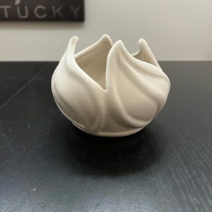 May include: A cream-colored, ceramic bowl with a unique, petal-like design. The bowl has a rounded base and the sides curve upwards, creating a flower-like shape. The interior is smooth, while the exterior has a textured, swirled pattern. The bowl sits on a dark surface.