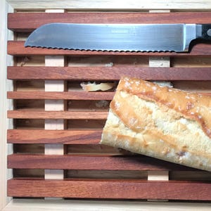 May include: A wooden bread cutting board with a stainless steel serrated knife and a partially sliced loaf of bread. The board is made of dark brown wood with a grid pattern. The bread is golden brown with a crusty exterior.