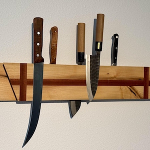 May include: A horizontal wooden magnetic knife strip featuring a light-toned wood grain with a dark wood inlay cross pattern. Five various kitchen knives with different blade shapes and handle materials are attached to the magnetic surface.