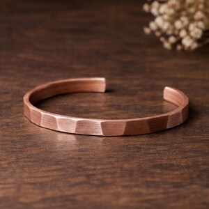 Op de afbeelding: Een koperen manchetarmband met een facetontwerp. De open armband heeft een warme, roodachtig bruine kleur en ligt op een donkere houten ondergrond. Het geometrische patroon van de armband geeft het een moderne, maar rustieke uitstraling.