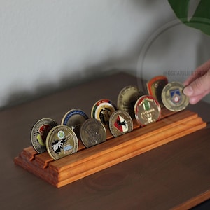 May include: A wooden coin display stand with multiple slots, holding various challenge coins. The stand is made of reddish-brown wood and is on a dark brown surface. A hand is placing a coin into the stand.
