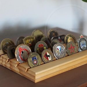 May include: A wooden display stand holding a collection of various coins. The stand is made of light-colored wood and features multiple slots to hold the coins upright. The coins have different designs and colors, with some featuring emblems and text.