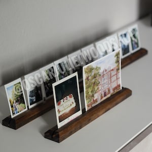 May include: Two wooden photo display stands holding several small framed photos. The stands are dark brown and have a slot to hold the photos upright. The photos depict various scenes, including a cake and a building.