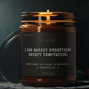 Puede incluir: Una vela encendida en un tarro de vidrio marrón con una etiqueta negra. La etiqueta presenta la cita "I CAN RESIST EVERYTHING EXCEPT TEMPTATION" de O. Wilde. La vela está hecha con cera de soja natural y es de Cultural Flume.