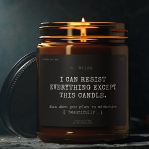 Puede incluir: Una vela encendida de cera de soja en un tarro de cristal marrón con una etiqueta negra. La etiqueta dice "I CAN RESIST EVERYTHING EXCEPT THIS CANDLE" de O. Wilde. La vela tiene una tapa dorada y el texto "Burn when you plan to misbehave beautifully."