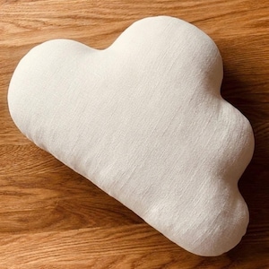 May include: A cloud-shaped throw pillow featuring a textured, off-white fabric surface. The pillow has a soft, rounded silhouette with multiple distinct lobes, mimicking the appearance of a cumulus cloud in a neutral, light cream color tone.