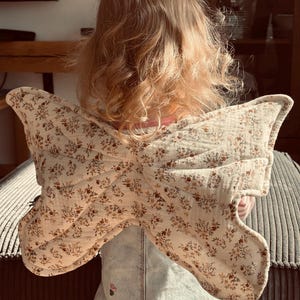 May include: Quilted fabric butterfly wings featuring a cream-colored base with a small-scale brown and orange floral print. The wings have a soft, padded texture and are shaped with distinct upper and lower lobes, designed to be worn on the back.