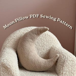 May include: A crescent moon-shaped pillow made of beige textured fabric, positioned in front of a cream-colored sherpa chair. The text "Moon Pillow PDF Sewing Pattern" is arched in white letters above the pillow against a dusty rose background.