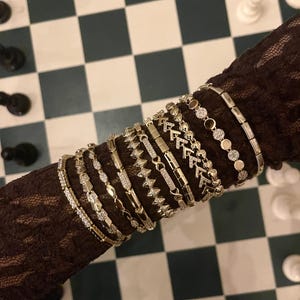 May include: A collection of gold-coloured bracelets with diverse designs, including geometric patterns and delicate embellishments. The bracelets are stacked on a dark, textured surface, creating a display of jewellery. The background features a chessboard.