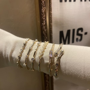 May include: Several gold-coloured bracelets, each with a unique design featuring geometric patterns and gemstones. The bracelets are presented on white fabric, highlighting their detailed craftsmanship. The background is neutral, allowing the jewellery to stand out.