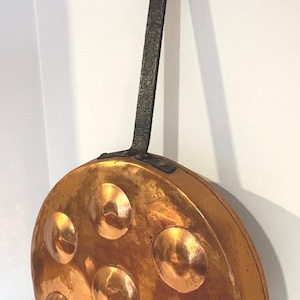 May include: Copper escargot pan with seven recessed wells. The pan has a long, textured, dark metal handle with a loop at the end for hanging. The pan's surface is a warm, reflective copper color.