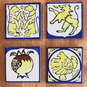 May include: Four square ceramic tiles with blue borders and white centers. Each tile features a different yellow illustration: two figures, a lion-like creature, a pomegranate, and a bird. The tiles are arranged on a wooden surface.