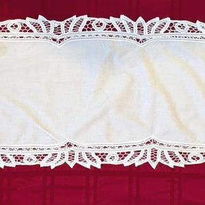 May include: A white rectangular table runner with intricate lace detailing along the edges. The lace features floral and geometric patterns. The runner is displayed on a red surface, creating a contrast.