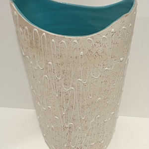 May include: A tall, curved ceramic vase with a bright turquoise interior. The exterior features a textured, off-white speckled finish overlaid with raised, swirling white lines that create a continuous, abstract pattern across the surface.
