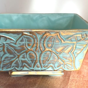 May include: A rectangular turquoise ceramic planter with gold trim and embossed leaf design. The interior is turquoise, and the exterior features a distressed gold finish. The planter sits on small feet.