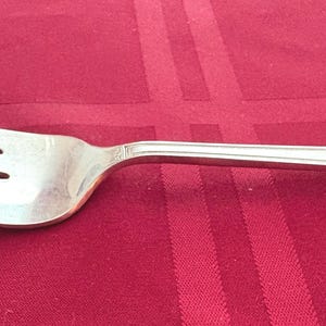 May include: A silver-colored metal dinner fork with four tines and a smooth, polished finish. The handle features a simple, straight design with a subtle beveled edge detail near the end, showcasing a clean and functional aesthetic for dining.