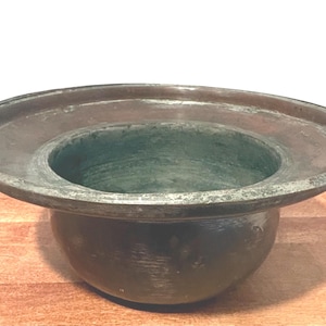 May include: A dark bronze-colored bowl with a wide, shallow rim and a rounded base. The bowl has a central, deeper well, and sits on a wooden surface. The metal has a weathered appearance.