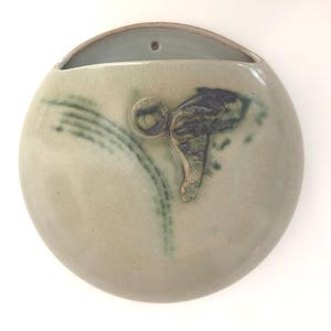 May include: A circular ceramic wall planter with a smooth, light gray glaze. The piece features a wide, open top opening and a small hole for mounting. A decorative accent of dark green and brown glaze creates a textured, abstract pattern.