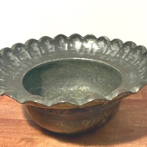 May include: A vintage metal bowl with a scalloped rim. The bowl has a dark, aged appearance with a textured surface. The bowl sits on a wooden surface. The bowl's design suggests it could be used for decorative or functional purposes.