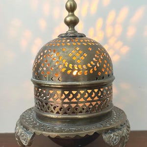May include: Ornate brass candle holder with a domed, perforated top and intricate base. The candle holder has a decorative finial and sits on three curved feet. The warm light from the candle shines through the cut-out patterns.