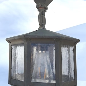 May include: An antique, hexagonal outdoor lantern with clear, textured glass panels and a dark metal frame. The lantern is suspended from a matching metal fixture and has a pyramidal top.