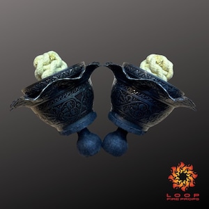 Può includere: Due oggetti di scena per il fuoco in metallo scuro ornato con motivi intricati e un materiale strutturato bianco sporco all'interno. Ogni oggetto di scena ha una base blu con una forma sferica. Il logo Loop Fire Props è visibile in basso a destra.