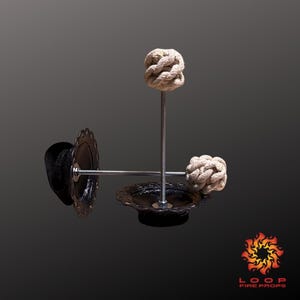 Puede incluir: Accesorio de fuego con base negra y bordes decorativos. Cuenta con una varilla metálica vertical central con una mecha anudada beige. Dos varillas horizontales se extienden desde la base, cada una con una mecha similar. Logotipo Loop Fire Props en la esquina inferior derecha.