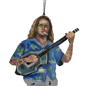May include: Wooden ornament of a person with long hair, sunglasses, and tattoos, playing a guitar. The person wears a blue Hawaiian shirt and white pants. The ornament hangs from a black string.