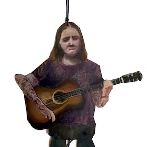 May include: Wooden ornament depicting a person playing an acoustic guitar. The figure wears a purple shirt and dark pants. The guitar is brown with a black fretboard. The ornament is suspended by a black string.